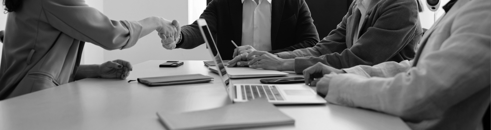 A picture of a table, laptop and notebooks, with people sitting around it and two shaking hands.
